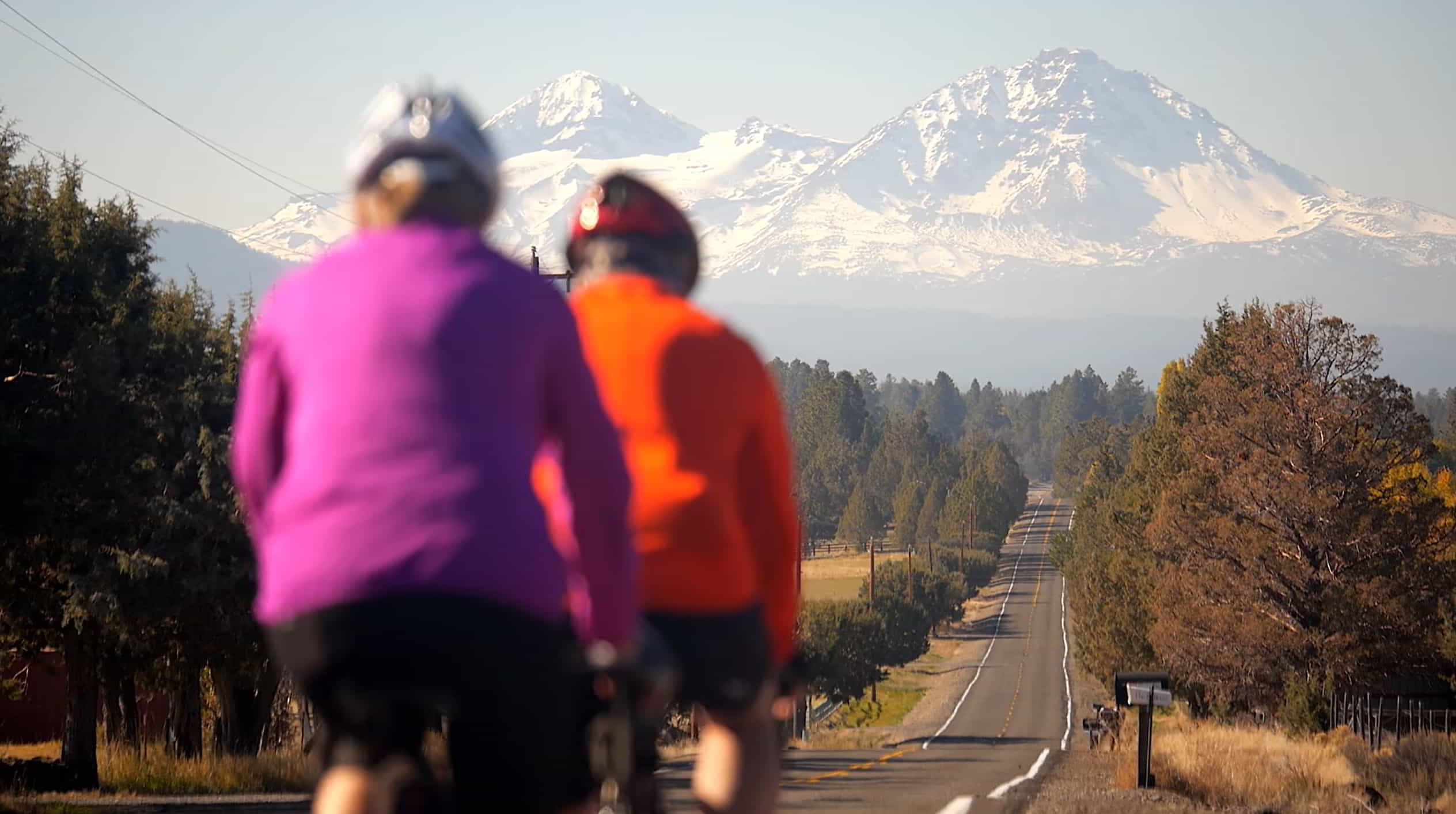 Close up of two bicyclist biking down a back road, away from the camera and towards the sisters mountains in the background